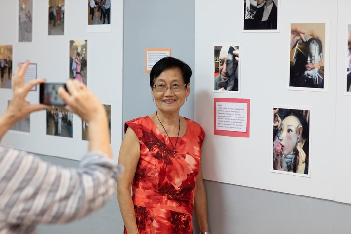 A photo of a woman standing in front of her photographer work. Another person is taking the artist's photo.