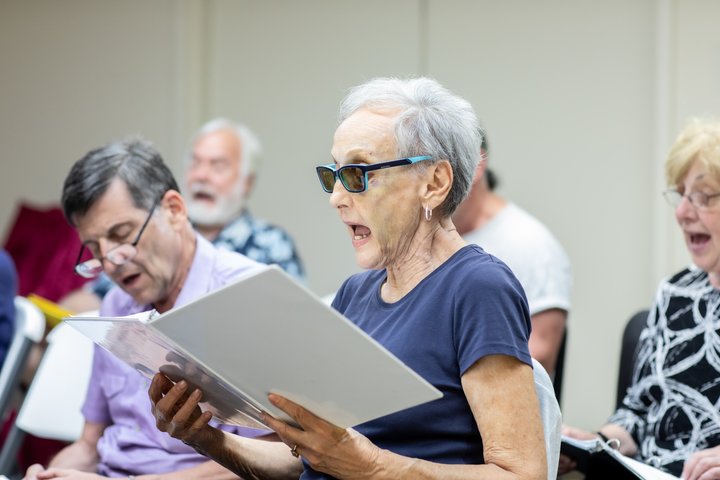 Several older adult choir members are standing and singing, holding sheet music.